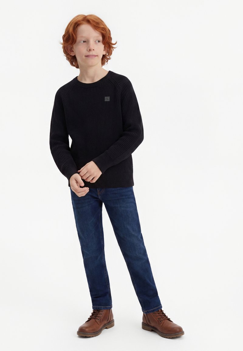 Jeune garçon roux debout, portant un pull noir, un jean bleu foncé et des bottes marron, regardant légèrement sur le côté.