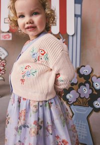 Jauns bērns ar cirtainiem blondiem matiem, ģērbies pasteļtoņu ziedu kleitā un rozā dzijas kardiganā ar izšūtiem ziediem, stāv telpās.