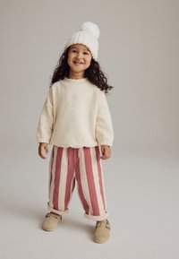 Un pull crème clair avec des poignets côtelés, associé à un pantalon rayé rose et blanc. Elle porte un bonnet tricoté avec un pompon et des chaussures beiges.