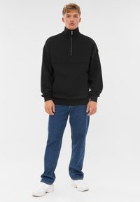 Felpa nera con zip e colletto alto, dotata di tasca a marsupio e polsini a costine, abbinata a jeans blue e sneakers bianche.