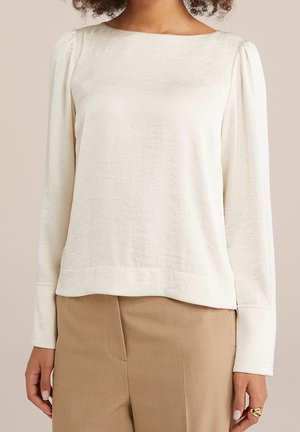 Nainen, joka pukeutunut valkoiseen pitkähihaseen puseroon, jossa on hienovarainen tekstuuri ja liehuvat olkapäät, yhdistettynä korkeavyötäröisiin beigen värisiin housuihin.