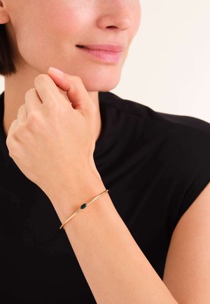 Gold bangle bracelet with a smooth, oval emerald accent. Worn on a wrist, featuring a minimalist design and sleek texture.