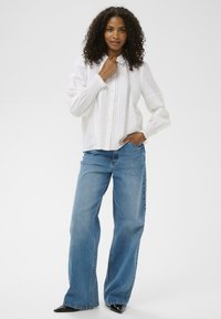 Femme avec des cheveux bouclés portant un blouse blanche à manches longues avec des détails en dentelle, un jean bleu ample, et des chaussures à talons pointus noires, se tenant avec une main dans sa poche.