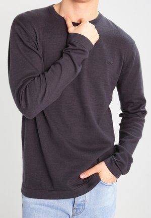 Osoba ubrana w ciemną bluzę z długim rękawem, trzymająca kołnierz jedną ręką, a drugą rękę ma wsuniętą do kieszeni jasnoniebieskich dżinsów.