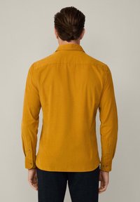 Camisa de manga larga amarilla mostaza hecha de una tela suave. Presenta un collar clásico, puños abotonados y una superficie lisa y texturizada.