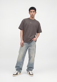 Junger Mann steht mit einer Hand in der Tasche, trägt ein dunkelgraues T-Shirt, locker sitzende ausgebleichte blaue Jeans und weiße Sneakers mit schwarzen Details.