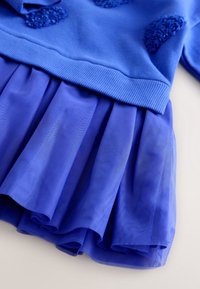 Robe bleue avec une ceinture côtelée et des détails floraux texturés sur le corsage, avec une jupe superposée plissée.