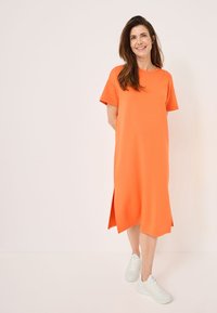 Orange short-sleeve dress with a straight cut, side slits, and a crew neckline, paired with white athletic shoes. Smooth fabric texture.
