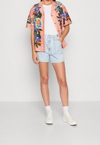 Persona vistiendo una camiseta blanca, una camisa de botones corta de mangas rosas con flores, pantalones cortos de mezclilla claros, calcetas blancas y zapatillas blancas, de pie contra un fondo simple.