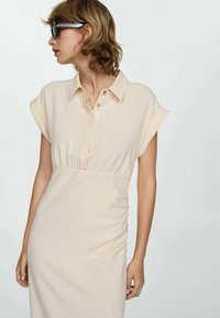 Vrouw met kort, golvend haar draagt een beige, geknoopte jurk met opgerolde korte mouwen en grote zwarte zonnebril, terwijl ze naar links kijkt tegen een effen achtergrond.