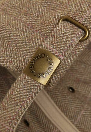 Nahaufnahme eines braunen Fischgrätstoff-Armbands mit einer Messingschließe, auf der "JOULES" in kreisförmigem Text eingraviert ist.