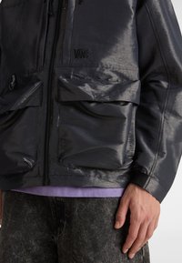 Gros plan sur une veste Vans noire brillante avec de grandes poches à rabat et une fermeture éclair, portée avec un jean foncé et une chemise violette en dessous.