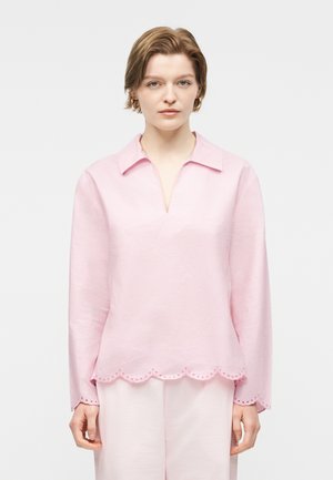 Mladá osoba s krátkymi svetlohnedými vlasmi, oblečená v svetloružový dlhý rukáv so zvlneným lemom a zodpovedajúcimi nohavicami, stojaca na bielom pozadí.