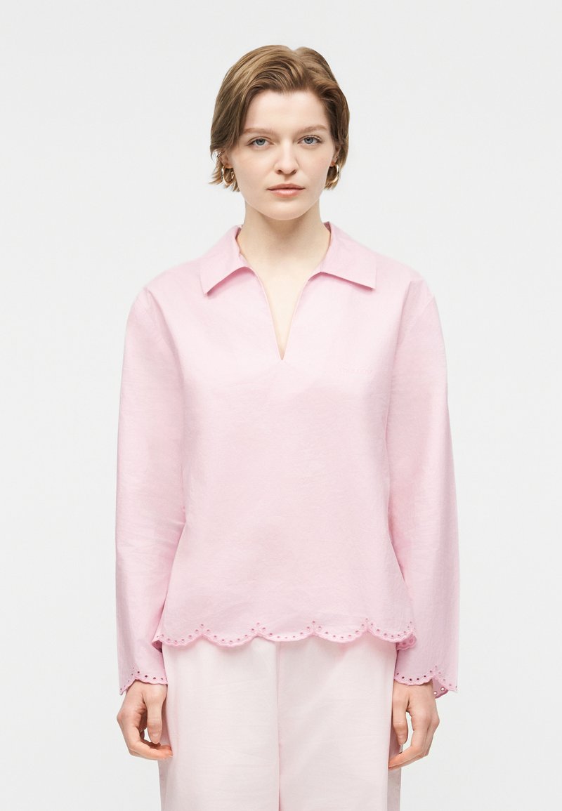 Fiatal személy rövid világosbarna hajjal, világos rózsaszín hosszú ujjú felsőt visel hullámos szegéllyel és hozzáillő nadrággal, fehér háttér előtt áll.