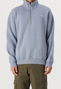 Light blue zip-up sweatshirt made of soft fabric, featuring a high collar, ribbed cuffs, and side pockets. Logo visible on the chest.