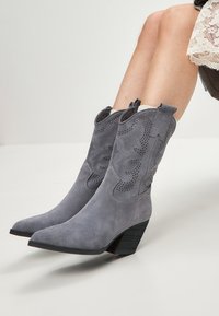 Boots de cowboy din suede gri, cu detalii perforate, vârf ascuțit și toc stratificat. Designul include bucle laterale pentru o utilizare ușoară.