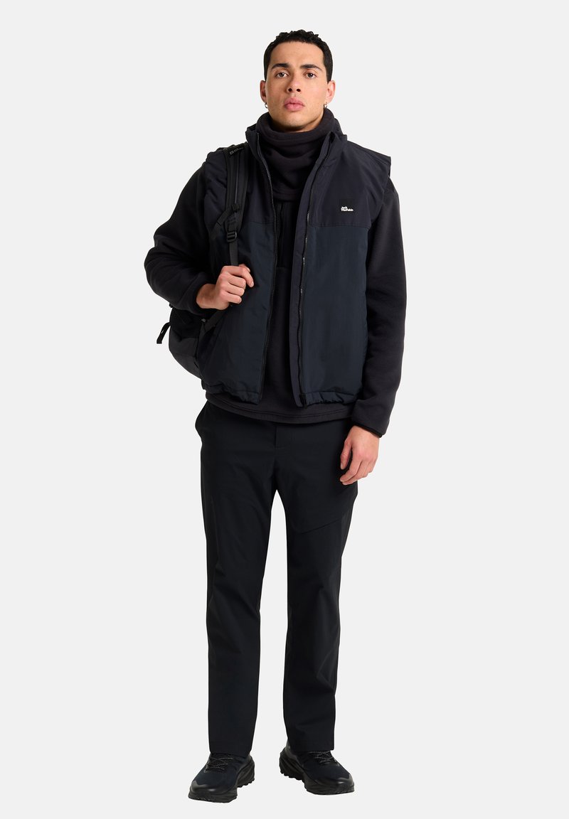 Giovane uomo in felpa nera, gilet blu navy, pantaloni neri e sneakers che tiene la tracolla di uno zaino nero, in piedi su uno sfondo bianco semplice.