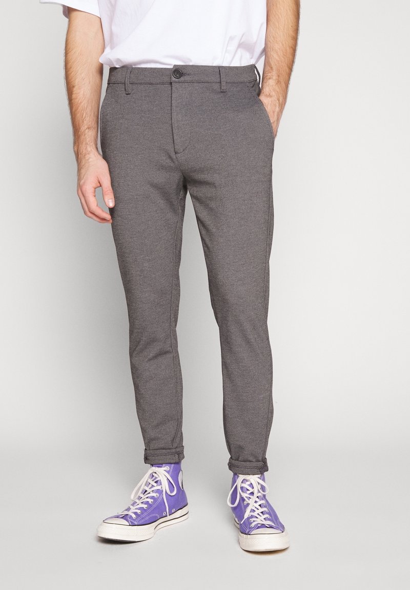 Homme portant un pantalon gris ajusté, un t-shirt blanc et des baskets montantes violettes, debout avec une main dans la poche.