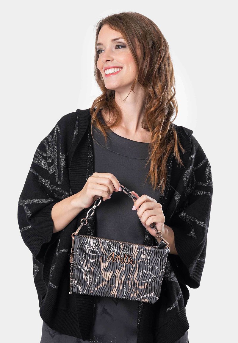 Bolso de mano a rayas negras y beige con una cadena de asa, que presenta una textura ondulada y un logo. La modelo sostiene el bolso mientras viste un atuendo negro.