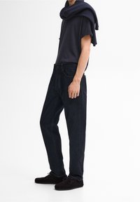 Mörkblå denimjeans med en avslappnad passform, tillsammans med en marinblå t-shirt och en ribbad stickad scarf som hänger över axlarna. Bruna mockaskor.