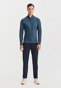 Granatowy sweter z suwakiem wykonany z miękkiego materiału, noszony z ciemnymi spodniami i białymi butami sportowymi z akcentami czerwieni i niebieskiego.