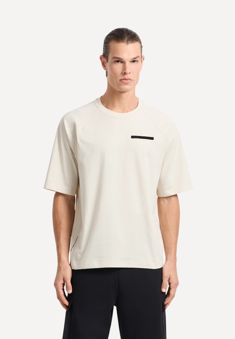 T-shirt corto a maniche corte di un beige chiaro con design raglan, caratterizzato da un piccolo accento rettangolare nero sul petto e dettagli discreti nelle cuciture laterali.