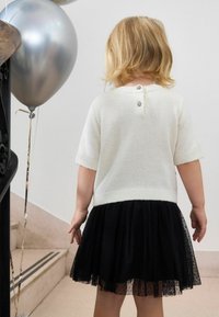 Ein kleines Kind mit blonden Haaren, das einen weißen Pullover und einen schwarzen Tüllrock trägt, steht nahe einer Treppe und hält einen silbernen Ballon.