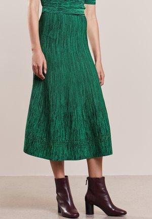 Femme portant une jupe midi texturée verte et des bottines en cuir marron, debout devant un fond beige uni.
