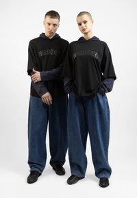 Două figuri poartă tricouri negre supradimensionate cu inscripția "WASTED" imprimată în gri, suprapuse peste mânecile lungi bleumarin. Acestea sunt acompaniate de pantaloni de blugi largi, de culoare albastră, și pantofi negri.