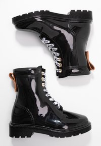 Bottines en cuir verni noir avec une semelle robuste, présentant des lacets décoratifs et une languette beige à l'arrière.