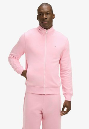 Homme portant une veste zippée rose clair et un pantalon assorti, debout avec une main dans la poche, expression neutre, fond blanc.