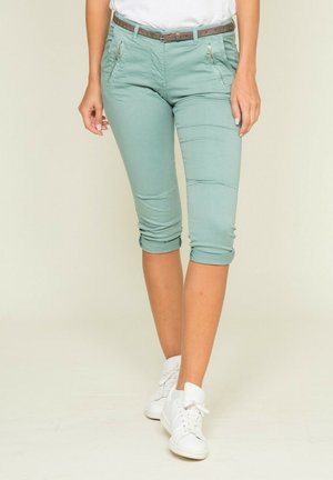Personne portant un pantalon court vert clair avec des poches zippées, une ceinture marron, des baskets blanches et un haut blanc sur un fond uni.