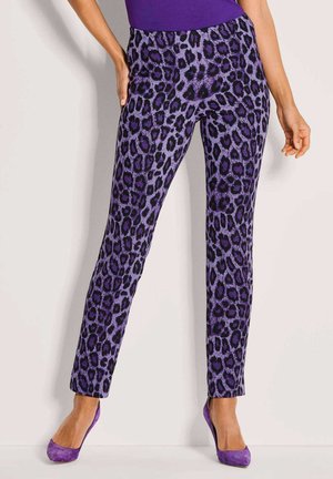 Vrouw in slim-fit paarse broek met luipaardprint en bijpassende paarse hoge hakken, staand tegen een effen lichte muur.
