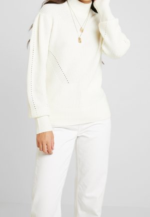 Femme portant un pull à col roulé côtelé blanc avec des détails en œillet et un pantalon blanc, accessoirisée avec deux colliers pendentifs dorés.