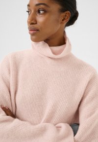 Svetlo roza turtleneck pulover iz mehkega, teksturiranega materiala, z ohlapnim krojem in širokimi rokavi, ki vključuje rebrast detajl na vratu.