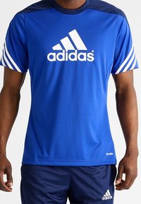 Sininen urheilupaita, jossa on valkoinen "adidas"-logo, lyhyet hihat ja raidoitetut yksityiskohdat hartioilla. Valmistettu kevyestä, kosteusihottumaa siirtävästä kankaasta.