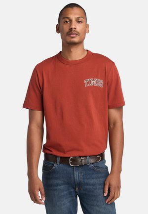 Röd t-shirt med kort ärm och en "TIMBS" grafisk design i vitt på vänster bröst. Bärs med blå denimjeans och ett brunt läderbälte.