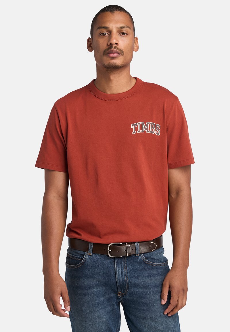 Red short-sleeve t-shirt with a "TIMBS" graphic in white on the left chest. Worn with blue denim jeans and a brown leather belt.