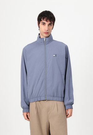 Chaqueta de color azul claro con cremallera y cuello alto, puños elásticos, dos bolsillos laterales y un pequeño logotipo en el pecho. Textura de tela suave.