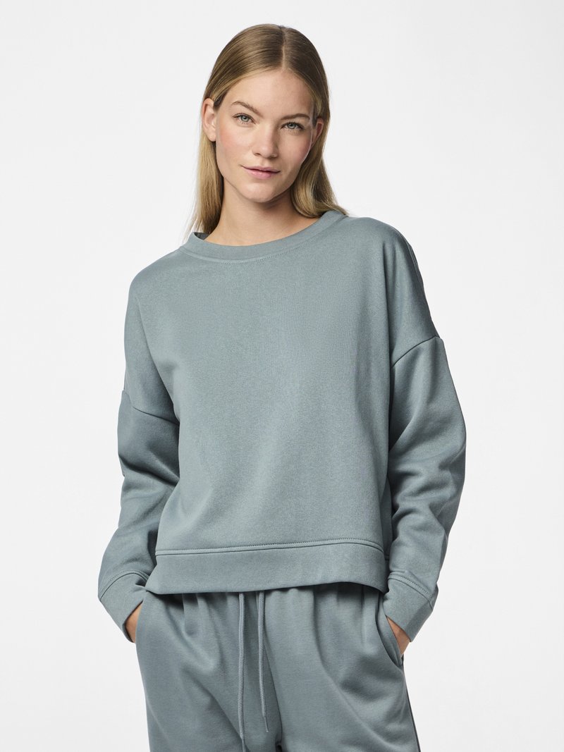 Kvinne med langt blondt hår iført lys blå hettegenser og matchende joggebukser, med hendene i lommene på hvit bakgrunn.