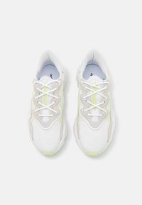 Zapatillas blancas y beige con material de malla, forma redondeada, cordones prominentes y acentos verdes a lo largo de los lados.