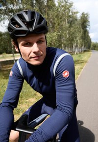 Velosipēdu apģērbs, kas ietver pieguļošu jūras zila garām piedurknēm topu ar atstarojošiem akcentiem un sarkanu zīmi, kombinēts ar melnu aerodinamisku ķiveri.