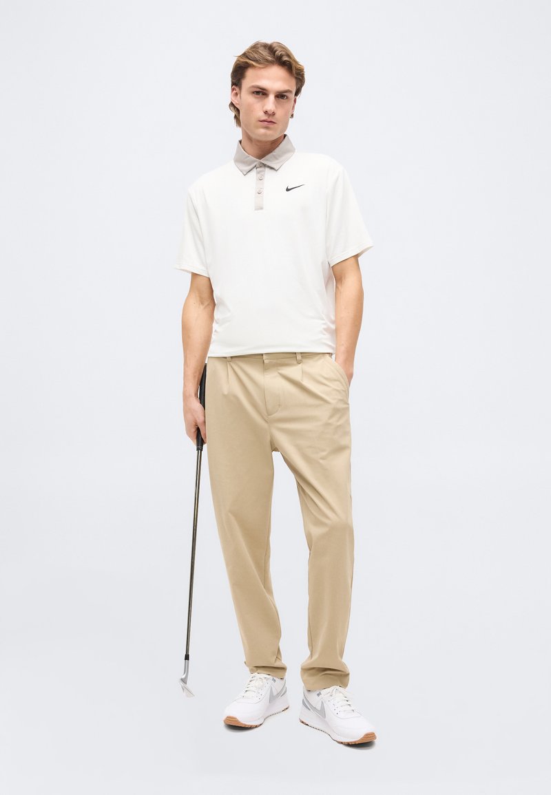 Jeune homme debout, une main dans la poche, tenant un club de golf, portant un polo blanc Nike, un pantalon beige et des baskets blanches.