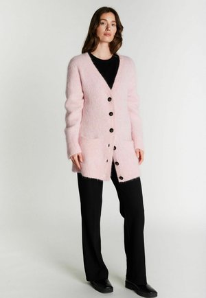 Femme portant un long cardigan duveteux rose avec des boutons noirs sur une chemise noire, un pantalon noir et des chaussures noires, debout devant un fond uni.