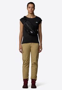 Černé přiléhavé tričko, béžové cargo kalhoty s kapsami a vínové turistické boty. Černá krosna je nošena přes hruď.