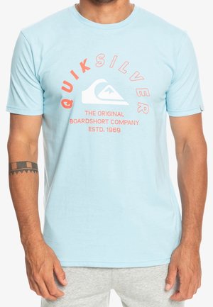 Mand iført en lyseblå Quiksilver t-shirt med koral-farvet tekst og logo, kombineret med lysegrå shorts, viser tatovering på venstre underarm.