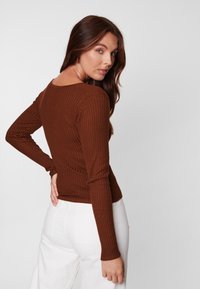 Haut à manches longues côtelé marron avec un décolleté en arrondi. Le tissu a une texture douce et est associé à un pantalon blanc taille haute.