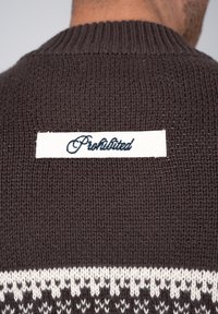 Gros plan d'un pull marron en tricot avec un patch blanc sur le haut du dos portant l'inscription "Prohibited" en écriture cursive.