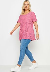 Túnica rosa con patrones florales cortados con láser, mangas cortas y escote redondeado, combinada con vaqueros ajustados de color azul claro y zapatillas blancas.