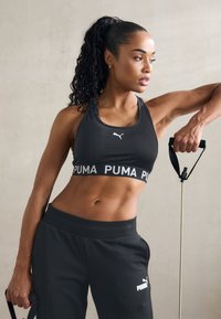 Crni sportski grudnjak s bijelim "PUMA" remenom, uparen s odgovarajućim crnim trenirkama. Tkanina izgleda glatko, a dizajn je prilagođen aktivnom odijelu.